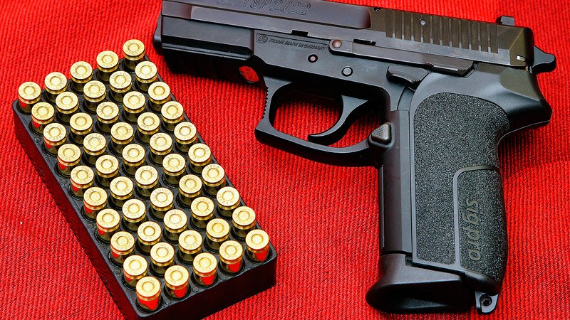 Black SIG Sauer SP2022 pistol beside a tray of brass handgun cartridges on a red background